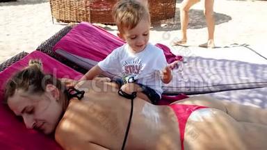 4k视频可爱的小男孩涂<strong>防紫外线</strong>乳液在母亲背上躺在海边的休息室